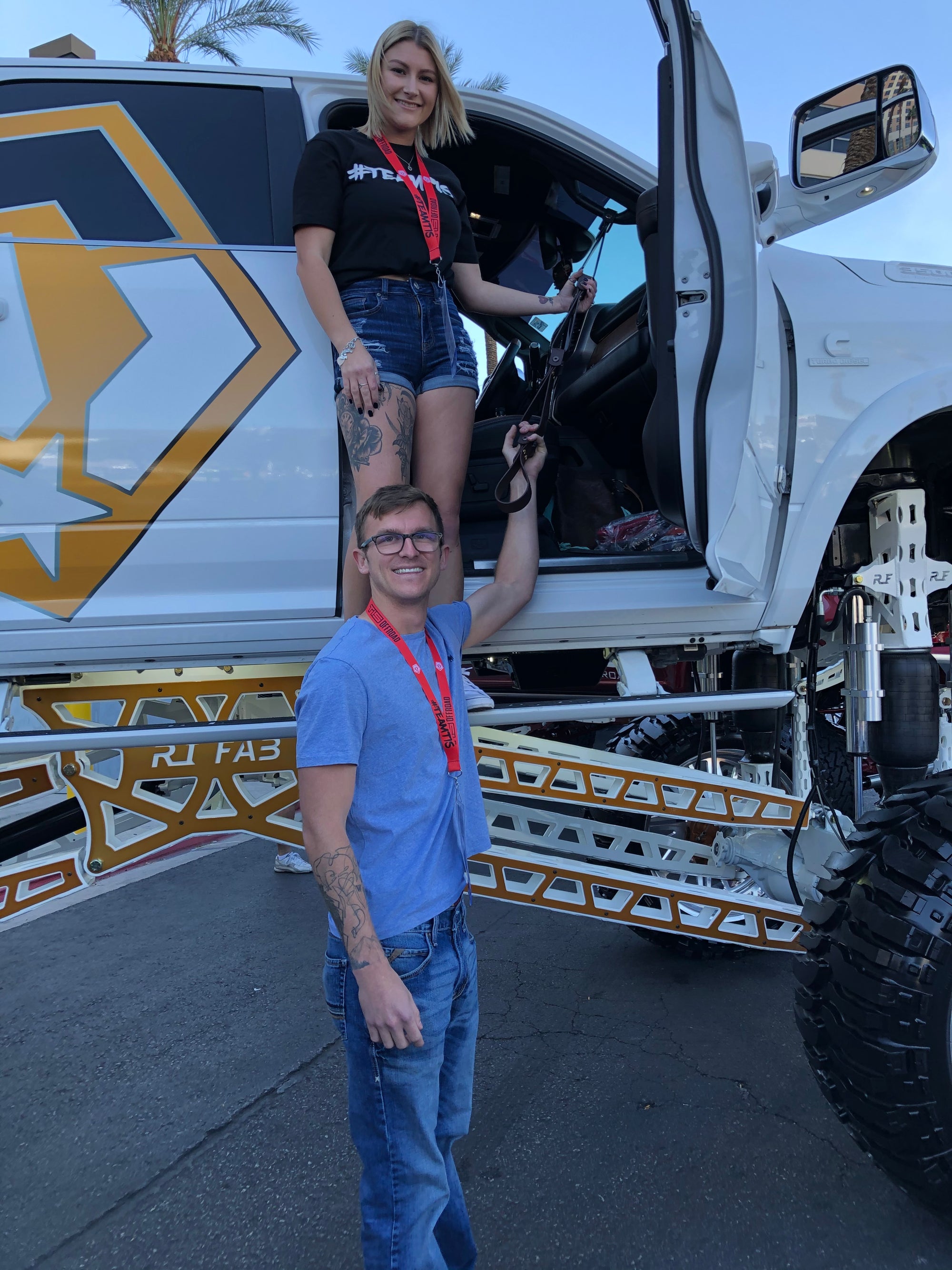 Two young people stand at the base and in the doorway of a very large lifted truck. Both hold extended grab handles meant for climb assistance