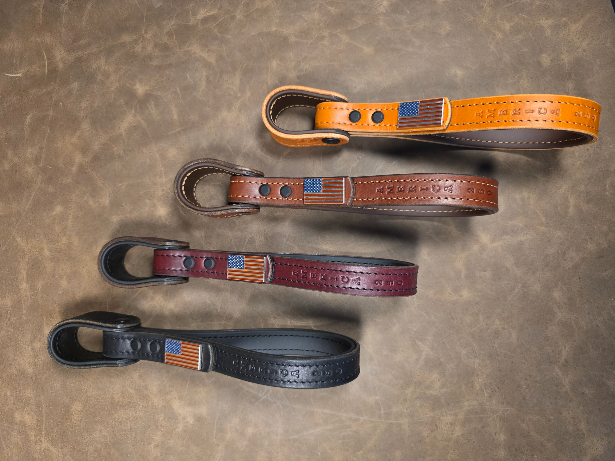 A series of truck entry assist grab handles in four different colors with Biothane core and tan stitching, enhanced with "America 250" stamping and an American Flag concho laying on a leather background.